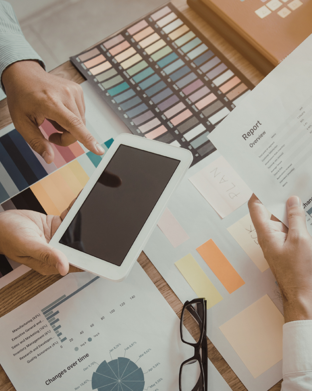 A group of people gathered around a table, reviewing reports, charts, color swatches, and a tablet, with a calculator visible.